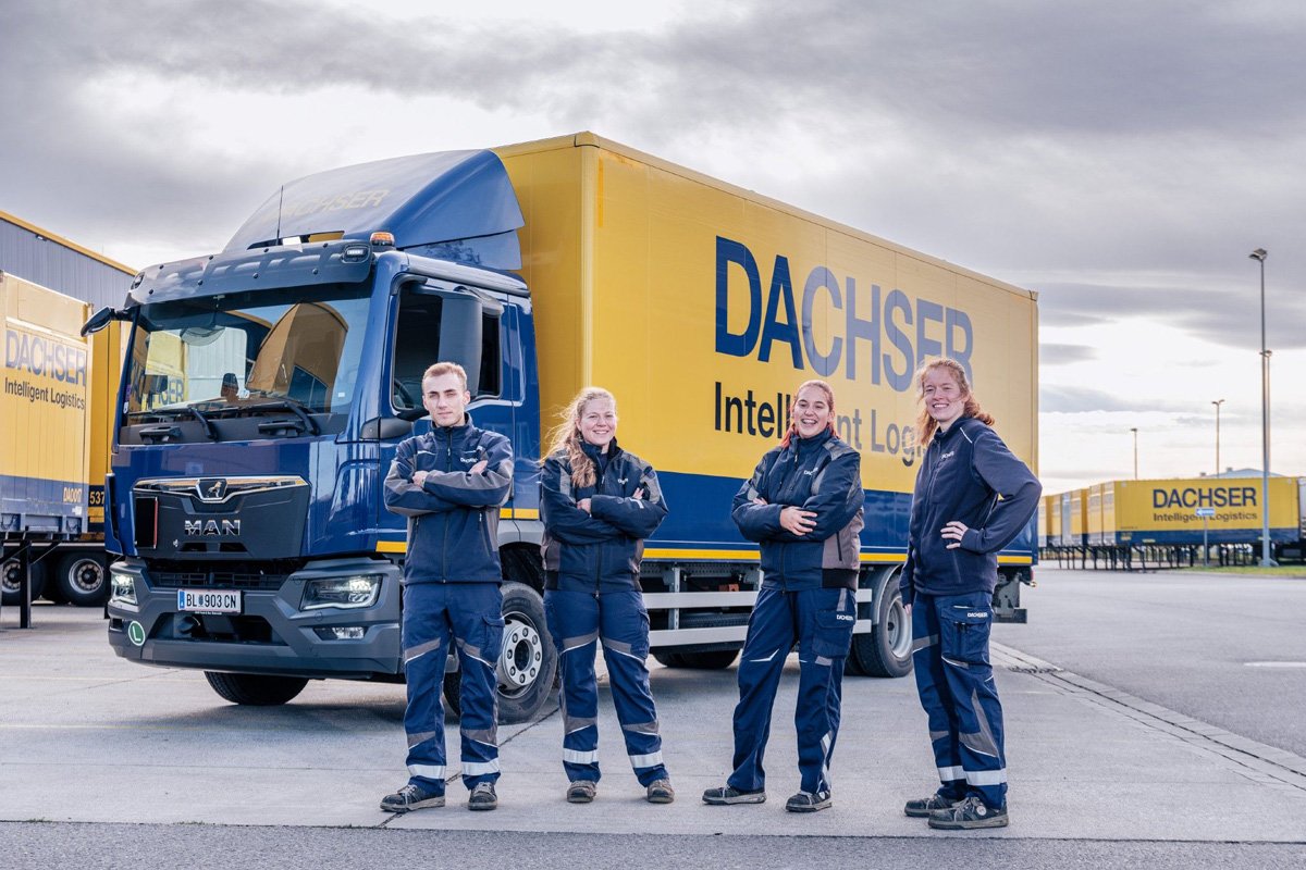 A group of four DACHSER employees stands confidently in front of a DACHSER branded truck. The truck has a blue cab and a yellow trailer with the company's logo and slogan "Intelligent Logistics" clearly visible. The employees are dressed in company uniforms, consisting of blue jackets and pants with reflective stripes. The background features additional DACHSER trailers and a partly cloudy sky.