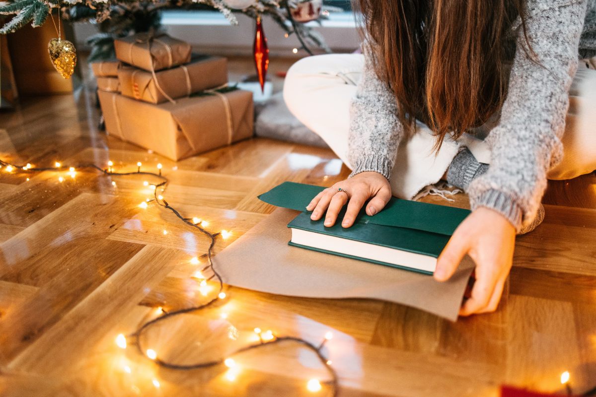 Una persona envolviendo un libro verde en papel marrón mientras está sentada en un suelo de madera, rodeada de luces navideñas brillantes y cajas de regalos bajo un árbol decorado.