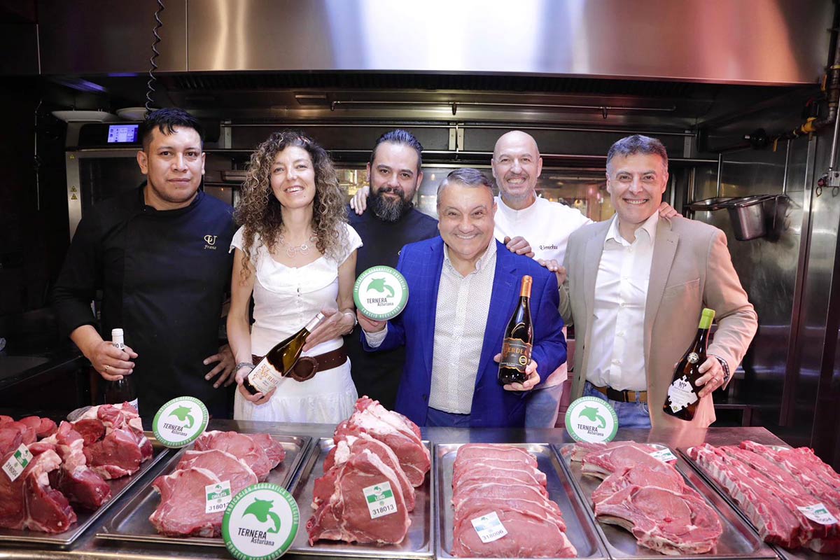 Seis personas posan en una cocina profesional detrás de bandejas con cortes de carne de vacuno, sosteniendo botellas de vino y material promocional de Ternera Asturiana en un evento gastronómico.