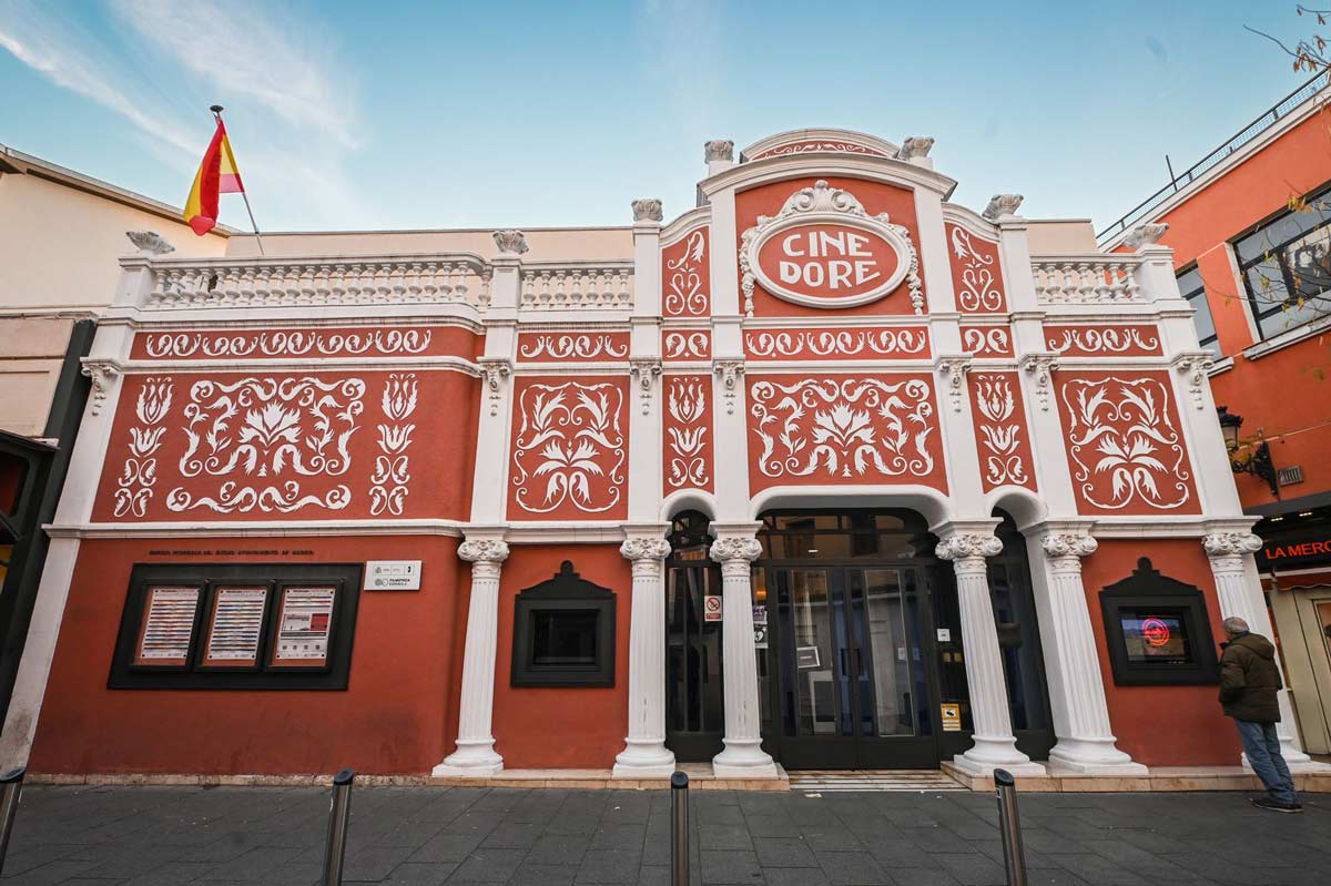 Fachada del Cine Doré, sede de la Filmoteca Española en Madrid. El edificio, de estilo modernista, presenta un frente rojo terracota decorado con motivos florales blancos en relieve, columnas estriadas y una balaustrada superior. Sobre la entrada, un medallón oval con el rótulo «CINE DORÉ». A la izquierda, vitrinas con la programación; en la cubierta ondea una bandera de España.
