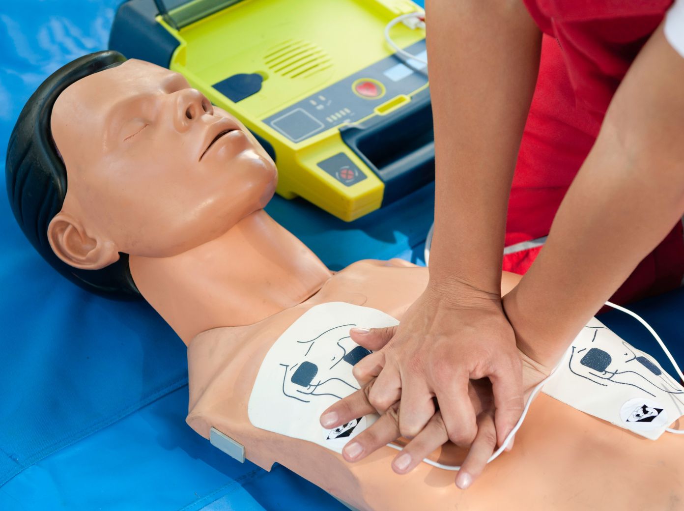 Persona realizando compresiones torácicas de reanimación cardiopulmonar (RCP) sobre un maniquí de entrenamiento con los electrodos de un desfibrilador externo automático (DEA) colocados en el pecho. El desfibrilador abierto se encuentra junto al maniquí durante una práctica de primeros auxilios.