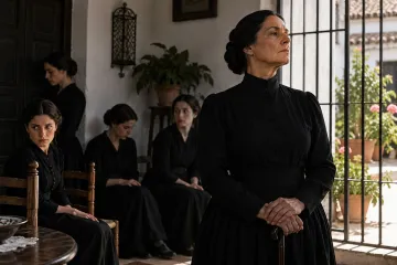 An older woman in mourning dress leaning on a walking stick beside a window grille, with four young women in black in the background of a whitewashed Andalusian courtyard
