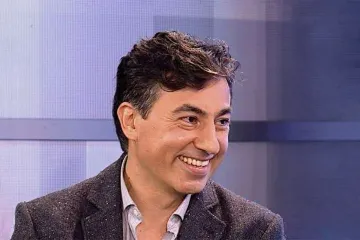 Portrait of baritone Marco Moncloa smiling, in a tweed blazer, in a television studio with a blue backdrop