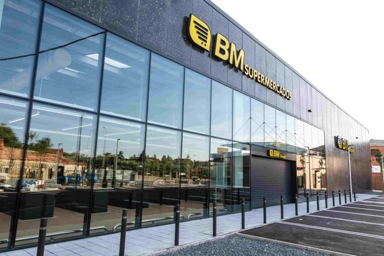 Fachada de un supermercado BM con un diseño moderno. La estructura del edificio presenta grandes ventanas de vidrio que reflejan el exterior, incluyendo árboles y edificios cercanos. El logotipo de BM Supermercados, en letras amarillas y blancas, es prominente en la parte superior de la fachada. El área del estacionamiento frente al supermercado está vacía.