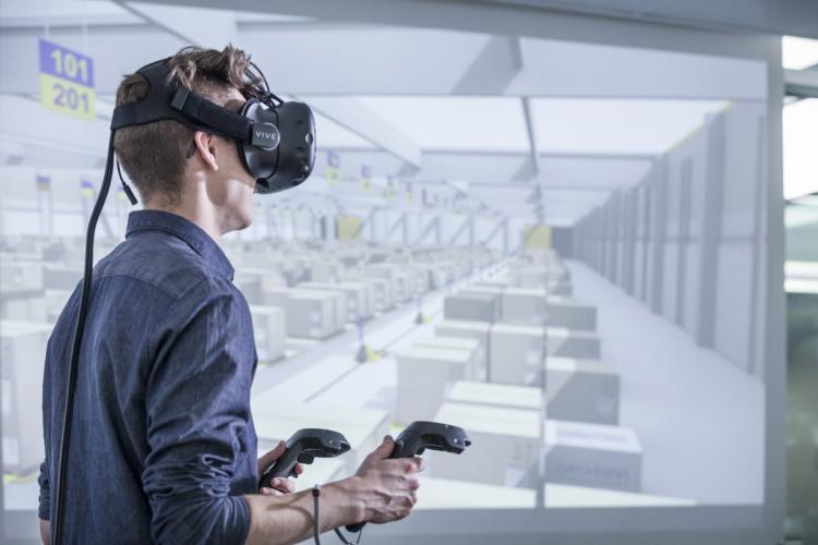 A man wearing a virtual reality headset and holding controllers explores a simulated warehouse environment displayed on a large screen in the background.