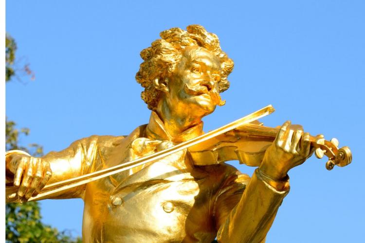 Primer plano de la estatua dorada de Johann Strauss II tocando el violín bajo un cielo azul brillante, mostrando los detalles intrincados de su rostro, cabello y violín.
