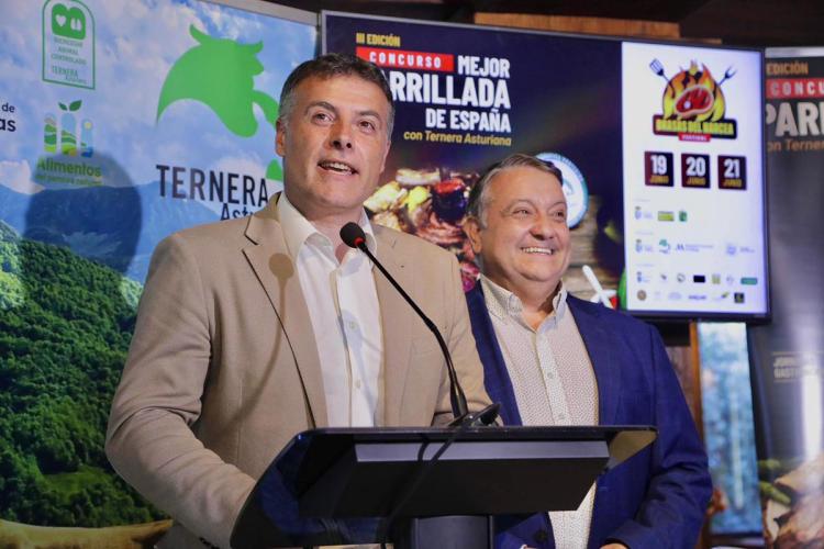 El alcalde de Brasas del Narcea habla desde un atril durante un evento promocional de Ternera Asturiana, con pantallas y carteles de un concurso de parrilla visibles al fondo