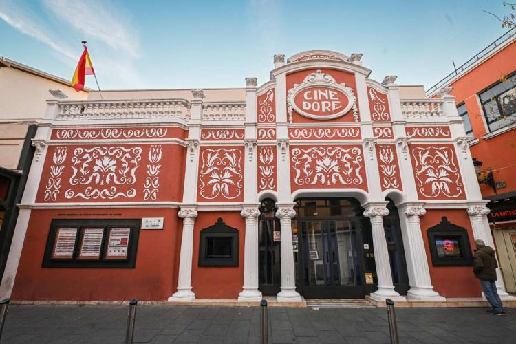 Fachada del Cine Doré, sede de la Filmoteca Española en Madrid. El edificio, de estilo modernista, presenta un frente rojo terracota decorado con motivos florales blancos en relieve, columnas estriadas y una balaustrada superior. Sobre la entrada, un medallón oval con el rótulo «CINE DORÉ». A la izquierda, vitrinas con la programación; en la cubierta ondea una bandera de España.