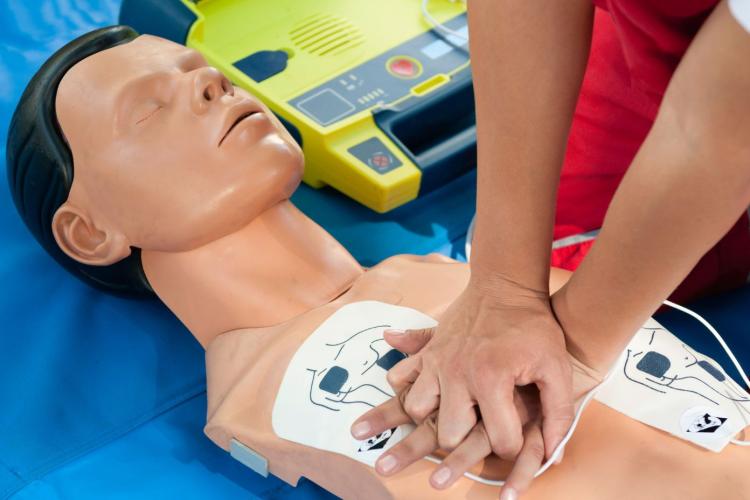 Persona realizando compresiones torácicas de reanimación cardiopulmonar (RCP) sobre un maniquí de entrenamiento con los electrodos de un desfibrilador externo automático (DEA) colocados en el pecho. El desfibrilador abierto se encuentra junto al maniquí durante una práctica de primeros auxilios.