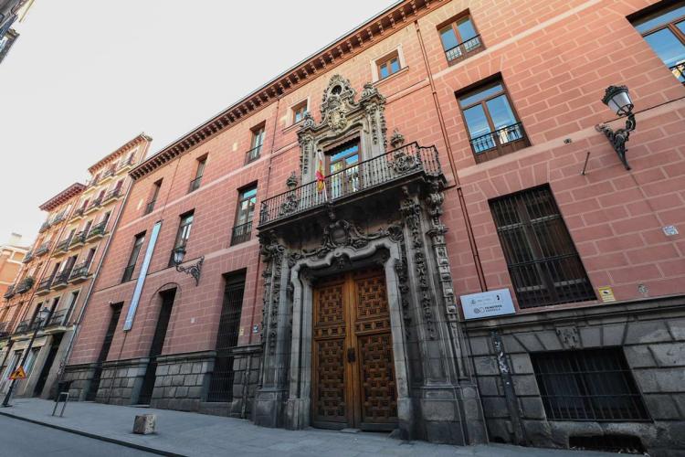 Fachada del Palacio del Marqués de Perales, sede de la Filmoteca Española en Madrid, vista en diagonal desde la calle. El muro, de color rosa salmón sobre un zócalo de granito, se abre en una portada barroca de piedra labrada con escudo heráldico y puerta de madera de dos hojas. Sobre ella, un balcón de forja con una bandera. A la derecha, el cartel de la Filmoteca Española.