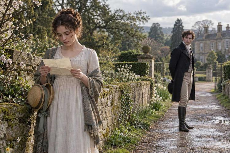 Woman in a Regency-era dress reading a letter beside a stone wall, with a man in a tailcoat watching from a country lane