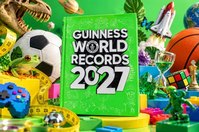 Green-covered Guinness World Records 2027 book surrounded by a football, a frog, a Rubik's cube, a rocket and other objects