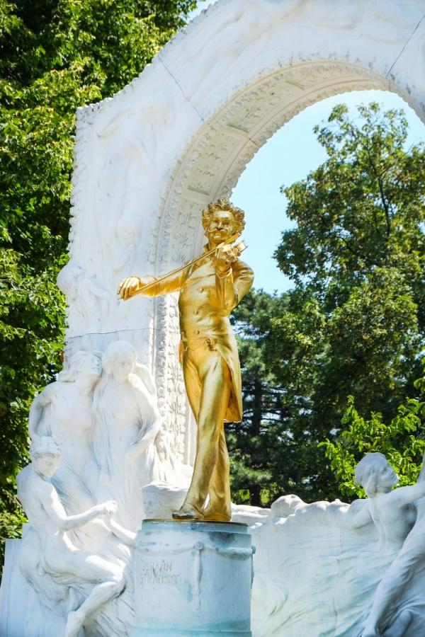 Vista completa del monumento a Johann Strauss II en Viena, con una estatua dorada rodeada por un arco de mármol blanco con esculturas ornamentadas, situado en un parque verde.