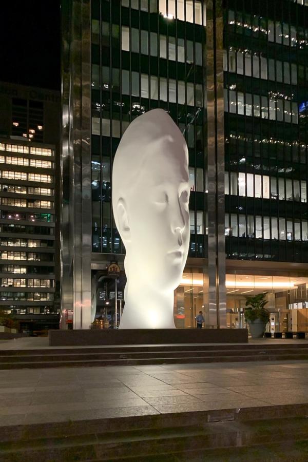 Soñando, gran escultura iluminada de una cabeza blanca serena de noche frente a un edificio moderno de cristal.