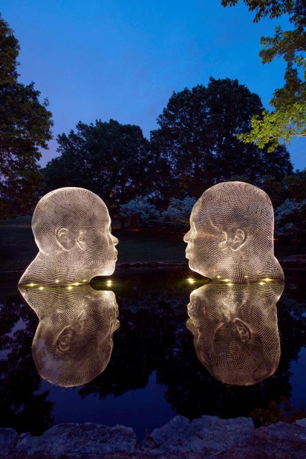 Awilda e Irma, de Jaume Plensa, dos grandes esculturas de cabezas de malla iluminadas que se miran frente a frente, reflejándose en el agua de noche con árboles al fondo.